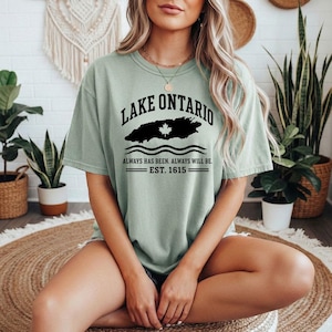 May include: A sage green short-sleeve t-shirt featuring black text that reads LAKE ONTARIO, a black silhouette of the lake with a white maple leaf, and the text ALWAYS HAS BEEN. ALWAYS WILL BE. EST. 1615 with horizontal lines above and below.