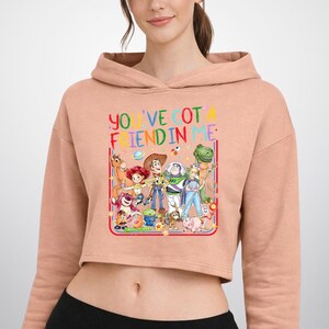 May include: A peach-colored cropped hoodie featuring a graphic print of various animated characters from Toy Story. The text "YOU'VE GOT A FRIEND IN ME" is printed in colorful letters above the characters, framed by a red and blue border.