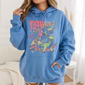 May include: A light blue hooded sweatshirt featuring a graphic print of Toy Story characters Woody, Buzz Lightyear, and Rex. The design includes the text "TOY story" and "SINCE 1995" with colorful geometric shapes in the background and a front kangaroo pocket.