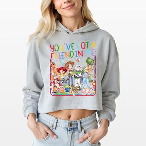 May include: A light gray cropped hoodie featuring a colorful graphic print of various animated toy characters. Above the characters, the text reads "YOU'VE GOT A FRIEND IN ME" in multi-colored, hand-drawn style lettering. The design is framed by a red border.