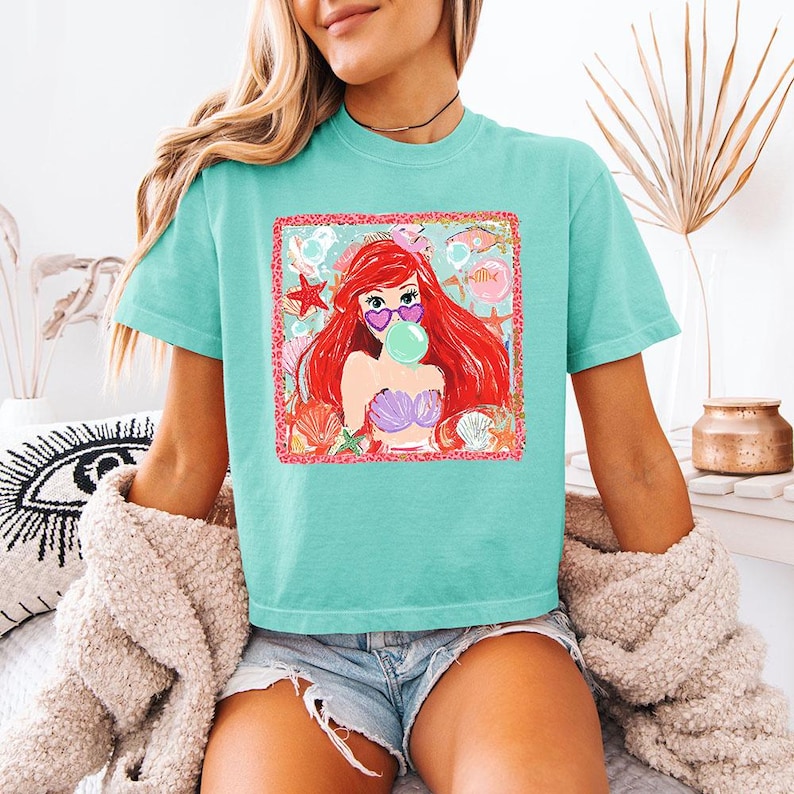 K&ouml;nnte beinhalten: Ein t&uuml;rkisfarbenes T-Shirt mit einer Grafik einer Meerjungfrau mit roten Haaren, herzf&ouml;rmiger Sonnenbrille und einer Blase. Die Meerjungfrau ist von Muscheln und Seesternen umgeben. Das Shirt ist kurz&auml;rmlig und hat einen Rundhalsausschnitt.