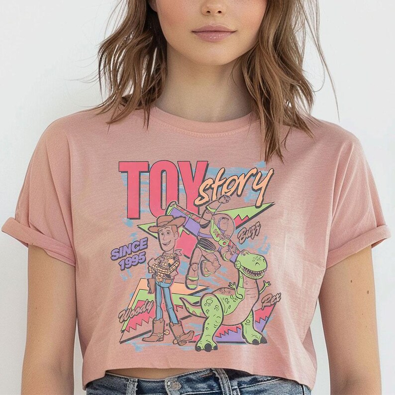 K&ouml;nnte beinhalten: Ein altrosa T-Shirt mit kurzen &Auml;rmeln und einem Grafikdruck der Toy Story-Figuren Woody, Buzz Lightyear und Rex. Das Design enth&auml;lt den Text "TOY story", "SINCE 1995", "Woody", "Buzz" und "Rex" vor einem Hintergrund aus bunten geometrischen Formen.