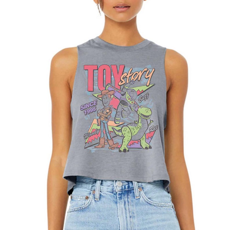 Puede incluir: Un top corto sin mangas de color gris jaspeado con un estampado gr&aacute;fico de Woody, Buzz Lightyear y Rex de Toy Story. El dise&ntilde;o incluye el texto "TOY story", "SINCE 1995", "Woody", "Buzz" y "Rex" con formas geom&eacute;tricas coloridas de fondo.