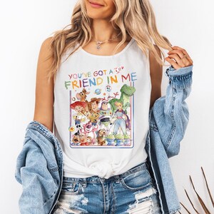 Pu&ograve; includere: Canotta bianca con una grafica colorata dei personaggi di Toy Story. Il testo "YOU'VE GOT A FRIEND IN ME" &egrave; visualizzato sopra i personaggi. Il design include Woody, Buzz Lightyear e altri personaggi.
