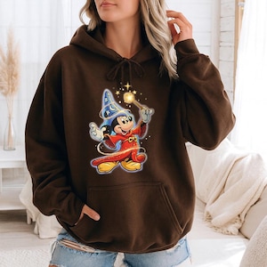 May include: A brown hooded sweatshirt featuring a graphic of Mickey Mouse dressed as a wizard, holding a wand with sparkling stars. The cartoon character is wearing a blue wizard hat and red robe. The sweatshirt has a front pocket and a drawstring hood.