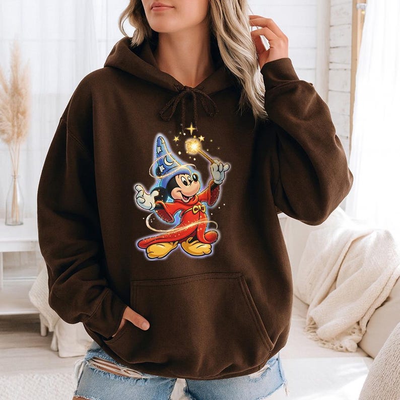 May include: A brown hooded sweatshirt featuring a graphic of Mickey Mouse dressed as a wizard, holding a wand with sparkling stars. The cartoon character is wearing a blue wizard hat and red robe. The sweatshirt has a front pocket and a drawstring hood.