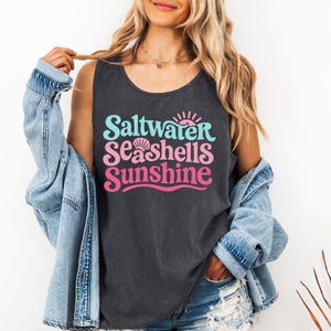 以下が含まれることがあります： ターコイズ、ピンク、マゼンタで「Saltwater, Seashells, Sunshine」という言葉が書かれた濃い灰色のタンクトップ。タンクトップはノースリーブで、ゆったりとしたフィット感があります。テキストには貝殻のグラフィックと太陽のグラフィックが含まれています。