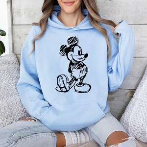 May include: A light blue hoodie featuring a black and white illustration of Mickey Mouse. The graphic depicts Mickey in a classic pose. The hoodie has a drawstring hood and a front pocket.