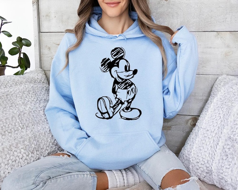 May include: A light blue hoodie featuring a black and white illustration of Mickey Mouse. The graphic depicts Mickey in a classic pose. The hoodie has a drawstring hood and a front pocket.