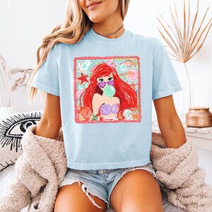 K&ouml;nnte beinhalten: Hellblaues T-Shirt mit einem Aufdruck einer rothaarigen Meerjungfrau, die eine herzf&ouml;rmige Sonnenbrille tr&auml;gt und eine Blase bl&auml;st. Die Grafik hat einen rosa Rahmen und Muschel-Akzente. Das T-Shirt hat kurze &Auml;rmel.