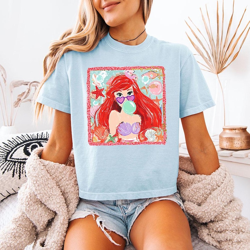 K&ouml;nnte beinhalten: Hellblaues T-Shirt mit einem Aufdruck einer rothaarigen Meerjungfrau, die eine herzf&ouml;rmige Sonnenbrille tr&auml;gt und eine Blase bl&auml;st. Die Grafik hat einen rosa Rahmen und Muschel-Akzente. Das T-Shirt hat kurze &Auml;rmel.
