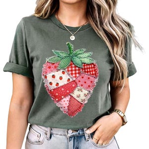 Puede incluir: Una camiseta verde con un diseño de fresa de patchwork. La fresa está compuesta por varias telas estampadas rojas y rosas, incluyendo cuadros, flores y corazones, con un tallo y hojas verdes. La camiseta tiene mangas enrolladas.