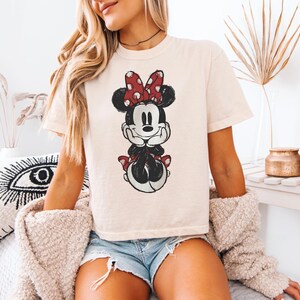 Puede incluir: Una camiseta de manga corta color crema con un estampado gr&aacute;fico desgastado de Minnie Mouse. El personaje aparece con su caracter&iacute;stico lazo rojo de lunares blancos, orejas negras y una expresi&oacute;n sonriente, centrado en la parte delantera.