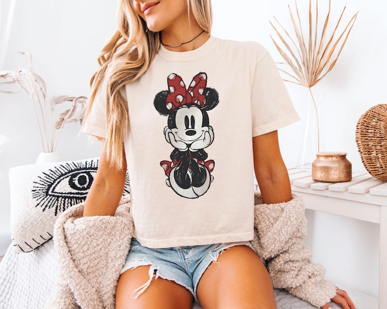 Puede incluir: Una camiseta de manga corta color crema con un estampado gr&aacute;fico desgastado de Minnie Mouse. El personaje aparece con su caracter&iacute;stico lazo rojo de lunares blancos, orejas negras y una expresi&oacute;n sonriente, centrado en la parte delantera.