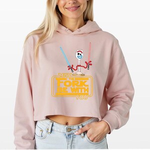 Puede incluir: Una sudadera corta de color rosa claro con un gr&aacute;fico del personaje Forky sosteniendo un sable de luz azul y otro rojo. Debajo del personaje, el texto amarillo "MAY THE FORK BE WITH YOU" aparece dentro de un dise&ntilde;o rectangular.