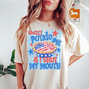 Op de afbeelding: Een crèmekleurig t-shirt met korte mouwen en een afbeelding van een taart met een Amerikaans vlagpatroon op de korst. De tekst SWEET POTATO PIE & I SHUT MY MOUTH staat in rode en blauwe letters, omringd door blauwe verticale strepen en sterren.