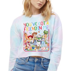 Peut inclure: Un sweat-shirt court &agrave; manches longues avec un motif tie-dye pastel, orn&eacute; d'une illustration color&eacute;e de divers personnages anim&eacute;s et du texte "YOU'VE GOT A FRIEND IN ME" en lettres arc-en-ciel. Le design inclut un cow-boy et un ranger.