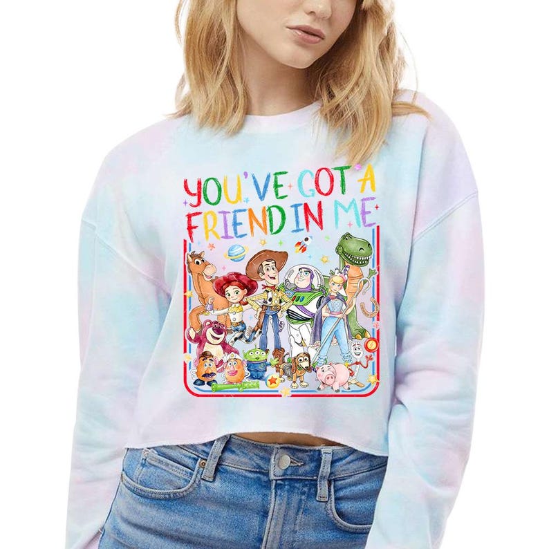 Peut inclure: Un sweat-shirt court &agrave; manches longues avec un motif tie-dye pastel, orn&eacute; d'une illustration color&eacute;e de divers personnages anim&eacute;s et du texte "YOU'VE GOT A FRIEND IN ME" en lettres arc-en-ciel. Le design inclut un cow-boy et un ranger.