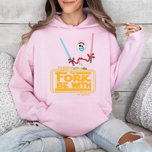 Op de afbeelding: Een lichtroze hoodie met een afbeelding van een witte spork met wiebeloogjes die een blauw en een rood lichtzwaard vasthoudt. Onder het figuur staat de gele tekst: MAY THE FORK BE WITH YOU, in een klassiek filmisch lettertype.