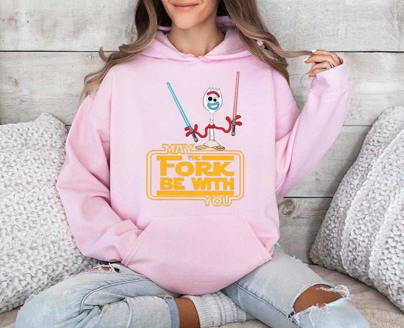 Op de afbeelding: Een lichtroze hoodie met een afbeelding van een witte spork met wiebeloogjes die een blauw en een rood lichtzwaard vasthoudt. Onder het figuur staat de gele tekst: MAY THE FORK BE WITH YOU, in een klassiek filmisch lettertype.