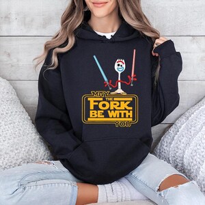 Op de afbeelding: Een zwarte hoodie met een afbeelding van een witte plastic vorkfiguur met rode pijpenrager-armen die een blauw en een rood lichtzwaard vasthoudt. Daaronder staat de gele tekst MAY THE FORK BE WITH YOU in een vetgedrukt, blokkerig lettertype.