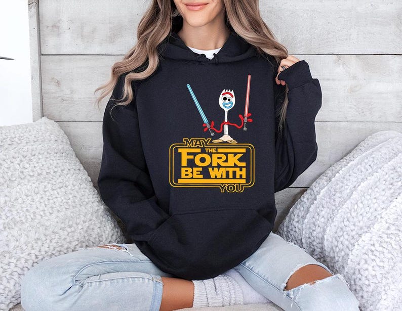 Op de afbeelding: Een zwarte hoodie met een afbeelding van een witte plastic vorkfiguur met rode pijpenrager-armen die een blauw en een rood lichtzwaard vasthoudt. Daaronder staat de gele tekst MAY THE FORK BE WITH YOU in een vetgedrukt, blokkerig lettertype.