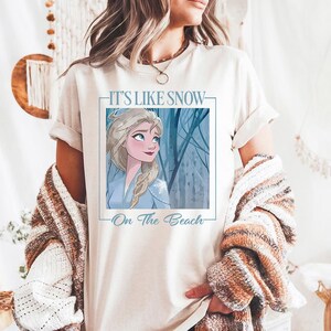 May include: White t-shirt with a graphic of Elsa from Frozen. The graphic says "It's Like Snow - On The Beach".