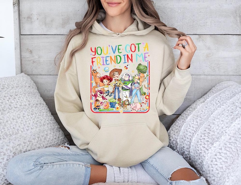May include: A beige hooded sweatshirt featuring a colorful graphic print of various animated characters from the Toy Story franchise. The text "YOU'VE GOT A FRIEND IN ME" is printed in multicolored letters above the group of characters.
