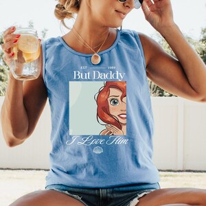 May include: A light blue tank top with the text "EST 1989 But Daddy I Love Him" and a graphic of a cartoon character with red hair. The tank top is sleeveless and made of a soft material.