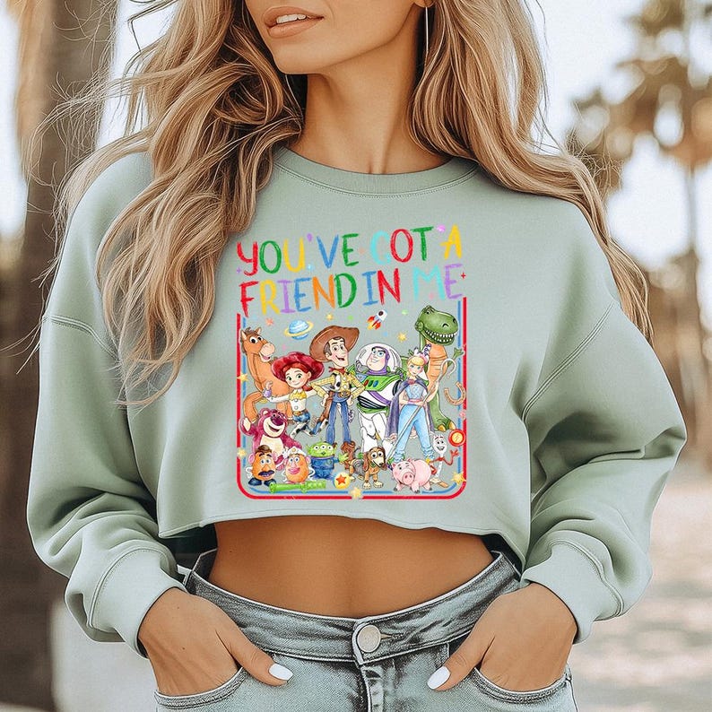 Peut inclure: Un sweat-shirt court &agrave; manches longues vert sauge avec une impression graphique color&eacute;e de divers personnages de Toy Story. Le texte "YOU'VE GOT A FRIEND IN ME" est imprim&eacute; en lettres multicolores au-dessus des personnages, entour&eacute; d'&eacute;toiles.