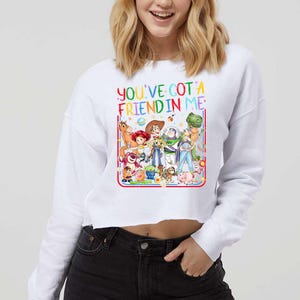 Peut inclure: Un sweat-shirt blanc &agrave; manches longues et coupe courte avec une impression graphique color&eacute;e repr&eacute;sentant divers personnages de jouets anim&eacute;s. Le texte "YOU'VE GOT A FRIEND IN ME" est imprim&eacute; en lettres multicolores au-dessus.