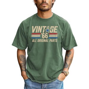 Puede incluir: Camiseta verde con el texto "VINTAGE '66 ALL ORIGINAL PARTS" en una fuente desgastada. El dise&ntilde;o incluye una estrella azul y rayas rojas y azules. La camiseta tiene mangas cortas y cuello redondo.