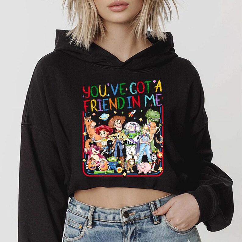 May include: A black cropped hoodie featuring a colorful graphic print of various animated characters from the Toy Story franchise. Above the characters, the text reads YOU'VE GOT A FRIEND IN ME in a vibrant, multi-colored, rainbow-style font.