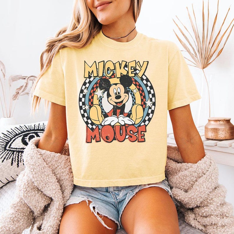 Pu&ograve; includere: Una t-shirt a maniche corte giallo pallido con una stampa grafica di Mickey Mouse centrata all'interno di una cornice circolare. Il design include il testo MICKEY MOUSE in lettere in grassetto, circondato da un bordo a scacchi, anelli colorati e stelle bianche.