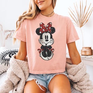 Puede incluir: Una camiseta de manga corta de color rosa claro con un estampado gr&aacute;fico desgastado de Minnie Mouse. El personaje aparece con su caracter&iacute;stico lazo rojo de lunares blancos, orejas negras y una sonrisa, apoyando la barbilla en sus manos.