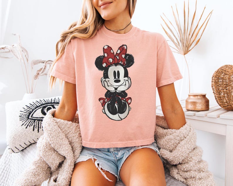 Puede incluir: Una camiseta de manga corta de color rosa claro con un estampado gr&aacute;fico desgastado de Minnie Mouse. El personaje aparece con su caracter&iacute;stico lazo rojo de lunares blancos, orejas negras y una sonrisa, apoyando la barbilla en sus manos.