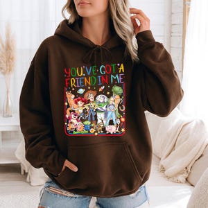 May include: A dark brown hooded sweatshirt featuring a colorful graphic print of various animated characters from the Toy Story franchise. The text "YOU'VE GOT A FRIEND IN ME" is printed in multi-colored letters above the character illustration.