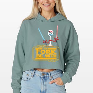 Puede incluir: Una sudadera corta de color verde salvia con un gr&aacute;fico de un personaje spork blanco con brazos rojos que sostiene un sable de luz azul y otro rojo. Debajo, el texto amarillo MAY THE FORK BE WITH YOU est&aacute; dentro de un marco rectangular.