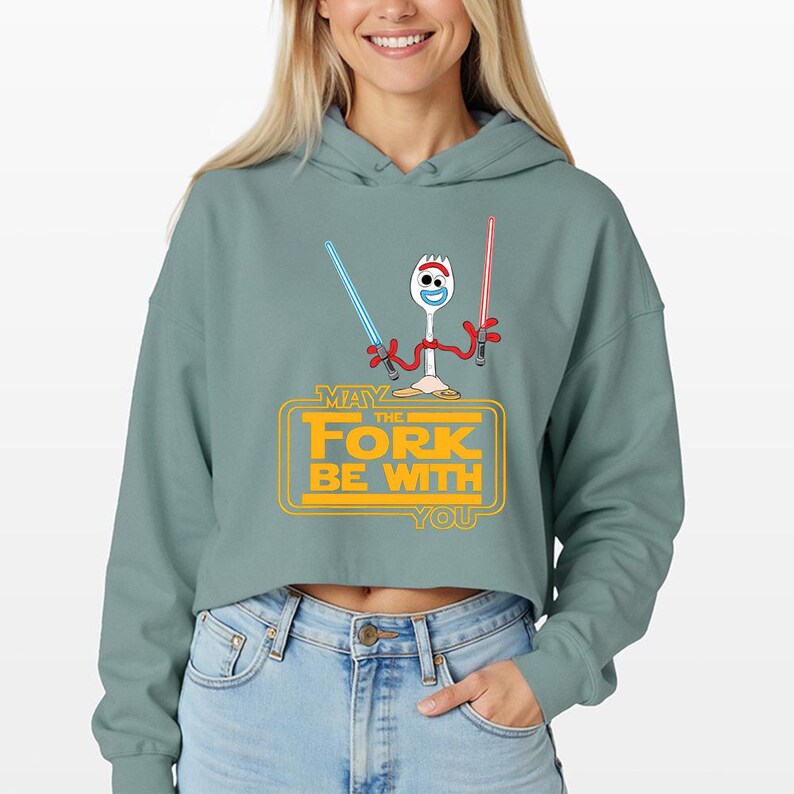 Puede incluir: Una sudadera corta de color verde salvia con un gr&aacute;fico de un personaje spork blanco con brazos rojos que sostiene un sable de luz azul y otro rojo. Debajo, el texto amarillo MAY THE FORK BE WITH YOU est&aacute; dentro de un marco rectangular.