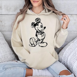 May include: A beige hoodie featuring a black and white sketch of Mickey Mouse. The hoodie has a kangaroo pocket and a drawstring hood. The graphic depicts Mickey Mouse in a classic pose, with his hands behind his back.