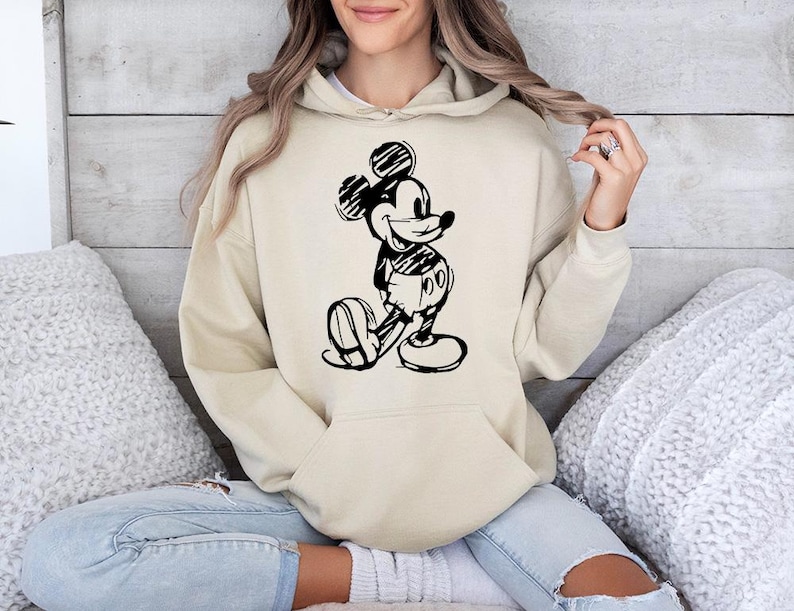 May include: A beige hoodie featuring a black and white sketch of Mickey Mouse. The hoodie has a kangaroo pocket and a drawstring hood. The graphic depicts Mickey Mouse in a classic pose, with his hands behind his back.
