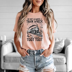 May include: Peach-colored t-shirt with a black and white graphic of a vintage train, desert landscape, and the text "I'M NOT ALWAYS A TRAIN WRECK JUST KIDDING TOOT TOOT". The shirt is made of soft material.