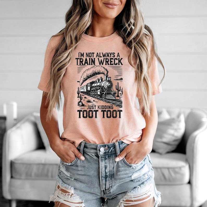 May include: Peach-colored t-shirt with a black and white graphic of a vintage train, desert landscape, and the text "I'M NOT ALWAYS A TRAIN WRECK JUST KIDDING TOOT TOOT". The shirt is made of soft material.