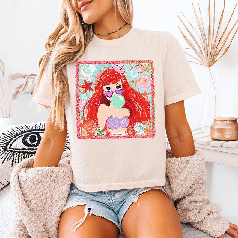 K&ouml;nnte beinhalten: Cremefarbenes T-Shirt mit einem farbenfrohen Aufdruck einer Meerjungfrau mit roten Haaren, die eine herzf&ouml;rmige Sonnenbrille tr&auml;gt und eine Blase bl&auml;st. Die Grafik ist mit einem rosa Rand umrahmt und enth&auml;lt Muscheln und Seesterne.