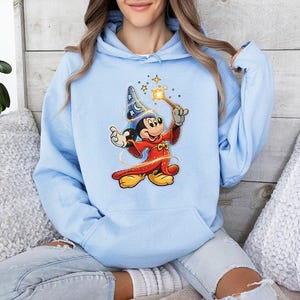 May include: Light blue hoodie featuring a graphic of Mickey Mouse dressed as a sorcerer, holding a wand with stars. The cartoon character is wearing a red robe, a blue hat, and yellow shoes. The hoodie has a drawstring hood and a kangaroo pocket.