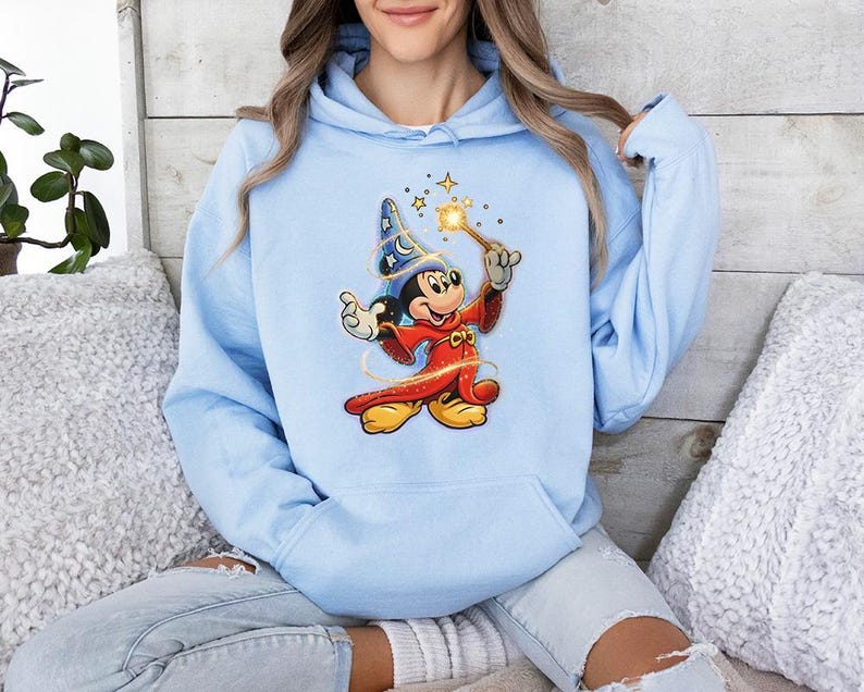 May include: Light blue hoodie featuring a graphic of Mickey Mouse dressed as a sorcerer, holding a wand with stars. The cartoon character is wearing a red robe, a blue hat, and yellow shoes. The hoodie has a drawstring hood and a kangaroo pocket.