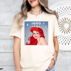 May include: A cream-coloured t-shirt with a square graphic of Ariel from The Little Mermaid. The graphic shows Ariel with red hair and blue eyes, the text "1989 Ariel's Version", and several seagulls. The t-shirt is a light, neutral colour.