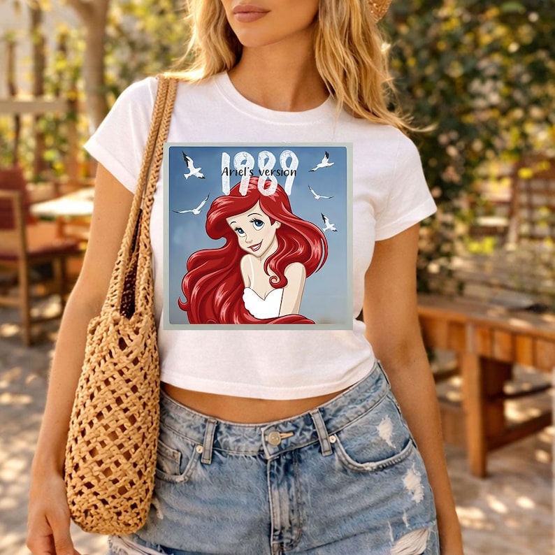 May include: White t-shirt featuring a square graphic of a cartoon mermaid with red hair, a white top, and the text "1989 Ariel's Version". The graphic includes seagulls and a blue background. The shirt is paired with denim shorts.