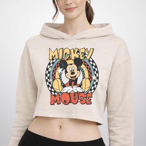 May include: A beige cropped hoodie featuring a graphic print of Mickey Mouse centered within a circular design. The graphic includes the text MICKEY MOUSE in yellow and red, surrounded by a black and white checkered border and white stars.