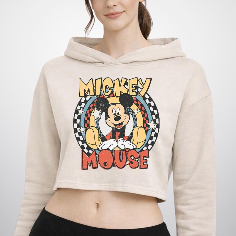 May include: A beige cropped hoodie featuring a graphic print of Mickey Mouse centered within a circular design. The graphic includes the text MICKEY MOUSE in yellow and red, surrounded by a black and white checkered border and white stars.